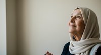 Mature woman wearing beige headscarf and dark clothing, looking upward thoughtfully against neutral background. Suitable for spirituality, faith, contemplation, and lifestyle content.