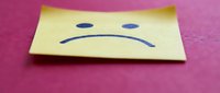 Yellow sticky note with sad face drawn in blue ink on pink surface expressing disappointment and negative emotions for mental health concepts.
