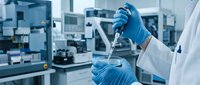 Scientist in blue gloves and white coat pipetting clear liquid in modern laboratory with advanced equipment and machinery in background.