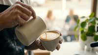 Barista pouring steamed milk into a latte art cup at a cozy cafe, creating a heart pattern in fresh espresso coffee.