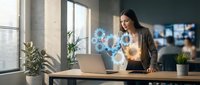 Young professional woman interacting with holographic digital interface at modern office workspace. Futuristic technology concept for business innovation.