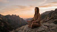 Young woman meditating in lotus position on mountain peak during golden hour sunrise, peaceful mindfulness practice in dramatic landscape setting.