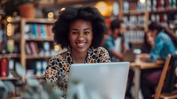 Young African American woman with curly hair smiling while working on laptop in cozy bookstore cafe with warm lighting and bookshelves in background.