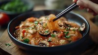 Glass noodles with mushrooms, vegetables and sesame seeds in black bowl. Chopsticks lifting steaming noodles, creating appetizing Asian cuisine atmosphere.