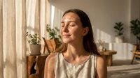 Young woman with long dark hair and light skin tone enjoying peaceful moment in bright, plant-filled interior space with natural sunlight streaming through curtains, conveying wellness and mindfulness.
