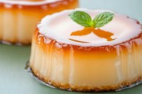 Creamy caramel flan dessert with mint leaf garnish on green surface, close up view showing smooth texture and golden caramel sauce dripping down sides.