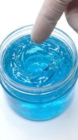 Hand touching bright blue gel or slime in clear container, creating ripple effect. Sensory play, stress relief, or craft activity concept for children and adults.