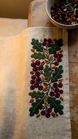 Embroidered linen table runner with cranberry and oak leaf pattern on rustic wooden surface, traditional folk art needlework for autumn home decor.