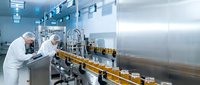 Workers in protective white uniforms operating automated bottling equipment in sterile pharmaceutical manufacturing facility with stainless steel machinery.