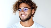 Young Hispanic man with curly hair and glasses smiling confidently against white background. Casual white t-shirt and modern eyewear showcase contemporary lifestyle.