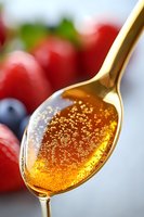 Golden honey dripping from spoon with tiny air bubbles creating sparkly texture, fresh berries in background for natural food photography and packaging design.