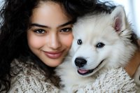 Young Asian woman in cozy sweater embracing fluffy white Samoyed puppy. Close-up portrait showing genuine connection between pet owner and dog companion.