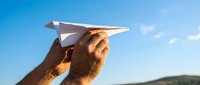 Hands holding white paper airplane against bright blue sky with clouds, symbolizing freedom, dreams, and new beginnings in outdoor setting.