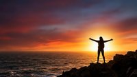 Person silhouette with arms raised celebrating freedom on rocky coastline at dramatic sunset over ocean waves with vibrant orange and purple sky colors.