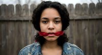 Young Hispanic woman with curly hair gagged with red rope in denim jacket against wooden fence background for awareness campaigns and social issues.