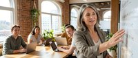 Mature woman with gray hair and light skin tone, wearing business casual attire, gestures while presenting to diverse colleagues in modern loft office with brick walls and large windows overlooking city skyline. Use for corporate training, business education, leadership, teamwork, and professional development materials.
