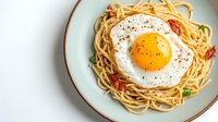Delicious spaghetti pasta topped with sunny side up fried egg, cherry tomatoes and fresh herbs served on white plate for breakfast or dinner meal.