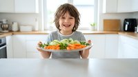 Happy young child with tousled brown hair proudly presenting colorful fresh vegetable salad plate in bright modern kitchen. Healthy eating lifestyle concept.