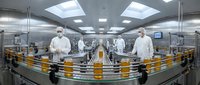 Pharmaceutical manufacturing facility with workers in protective suits monitoring automated production line with orange bottles in sterile cleanroom environment.