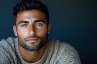 Close up portrait of young Mediterranean male model with dark hair and beard wearing beige sweater against dark background, looking directly at camera.