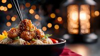 Steaming Asian noodles topped with glazed sesame chicken in dark bowl against blurred bokeh lights background. Garnished with chili and green onions.