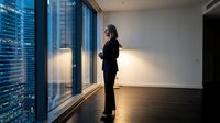 Professional businesswoman standing by floor-to-ceiling windows overlooking illuminated city skyline at night in modern office space with hardwood floors.