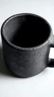 Rustic black ceramic mug with rough textured surface on white background, handcrafted stoneware cup for coffee or tea, minimalist style.