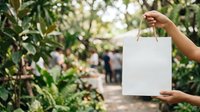 Person holding blank white shopping bag in botanical garden with lush green plants and foliage background, suitable for retail and eco-friendly branding.