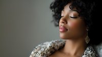 Young African American woman with natural curly hair and gold earrings in peaceful meditation pose against soft background, eyes closed expressing serenity and mindfulness.