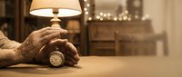 Elderly hands holding vintage pocket watch on wooden table in warm cozy living room with lamp light creating nostalgic atmosphere for memories and time concept.