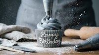 Dark charcoal birthday cupcake being frosted with piping bag, flour dust flying, rustic bakery setting, celebration dessert decoration.