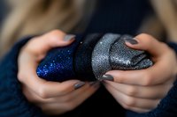 Female hands with dark gray manicure holding glittery blue and silver decorative ornament against dark background, creating magical holiday atmosphere.