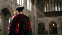 Graduate in academic regalia standing in gothic university hall during commencement ceremony, celebrating educational achievement and academic success.