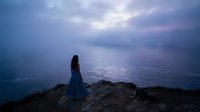 Young woman in flowing blue dress standing on rocky cliff overlooking calm ocean at twilight with dramatic purple and pink sky clouds creating peaceful solitude scene.