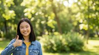 Young Asian woman wearing denim shirt giving thumbs up gesture in sunny park with green foliage background, expressing positivity and approval.