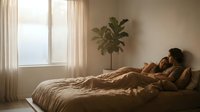 Woman with dark hair and medium skin tone resting on bed in warm, sunlit bedroom with potted plant and sheer curtains, conveying relaxation and comfort.