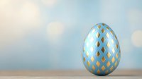 Decorative Easter egg painted in light blue and gold geometric diamond pattern standing on wooden surface against soft blurred bokeh background.