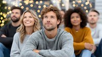 Young Caucasian man in gray sweater looking up thoughtfully with diverse group of friends behind him against warm bokeh lights background.