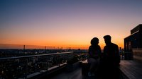 Two people standing on a rooftop terrace overlooking an urban skyline at sunset, silhouetted against an orange and purple sky with city lights below.