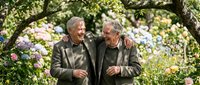 Two happy senior Caucasian men walking together in blooming garden, embracing friendship and enjoying retirement lifestyle outdoors among colorful flowers.