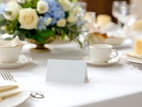 Elegant table setting with blank place card, white teacups, and floral centerpiece featuring blue and cream roses on crisp white tablecloth for wedding or event.