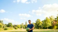 Young man with glasses holding a watermelon in an open field surrounded by green trees and blue sky, suitable for summer lifestyle and healthy eating content.