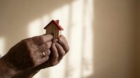 Hands gently holding small wooden house model with red roof in warm sunlight, symbolizing home protection, real estate investment and property care.