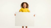 Young child with curly reddish-brown hair wearing bright yellow shirt holds blank white poster board against neutral background, ready for custom text or design messaging.