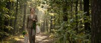 Young woman in beige coat walking peaceful forest path with golden sunlight filtering through green trees creating serene natural atmosphere for wellness content.