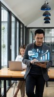 Asian businessman analyzing digital financial data and growth charts in modern office with colleague working in background.