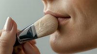 Close-up of makeup brush applying powder foundation on female face, showing professional cosmetic application technique with soft beige tones and natural lighting.