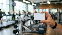 Man holding blank white business card in modern gym facility with blurred fitness equipment and people exercising in background for corporate branding and membership promotion.