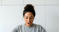 Young woman with dark hair styled in a top bun, medium skin tone, wearing casual gray t-shirt, smiling with mouth open in joyful expression against white tiled wall background.