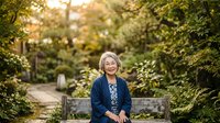 Senior woman with gray hair and fair complexion, wearing a navy cardigan and floral blouse, smiling while seated on a wooden bench in a lush garden setting surrounded by green foliage.